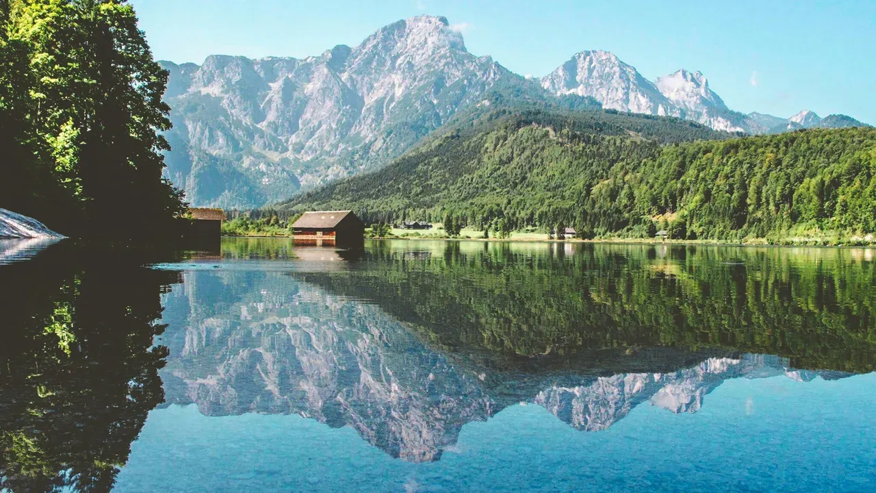 A calm alpine lake mirroring wooded slopes and snow-flecked rocky peaks in the Austrian Alps, with a wooden boathouse on the far shore