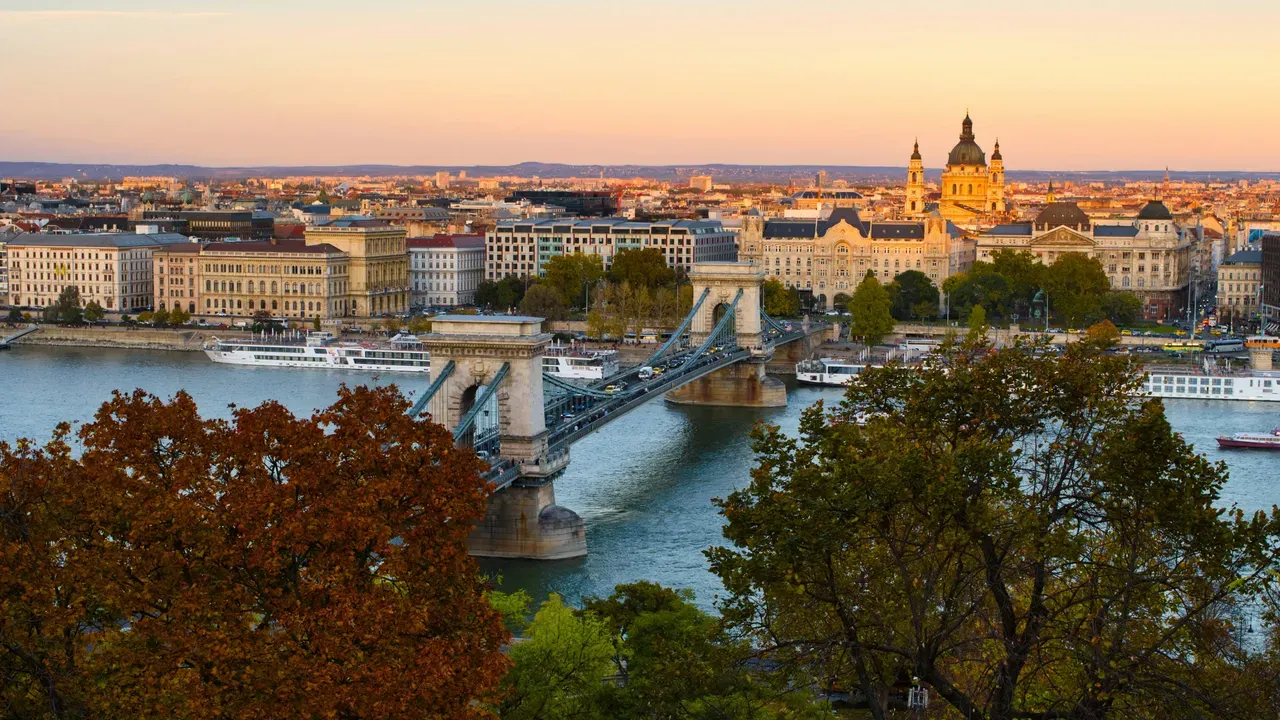 The Széchenyi Chain Bridge over the Danube at dusk, with St. Stephen's Basilica and the Budapest skyline behind, framed by autumn trees