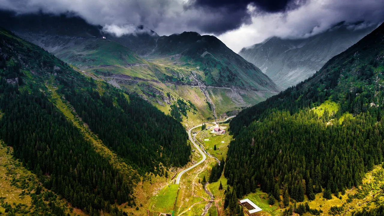 A mountain road winding through a deep forested Carpathian valley under heavy storm clouds