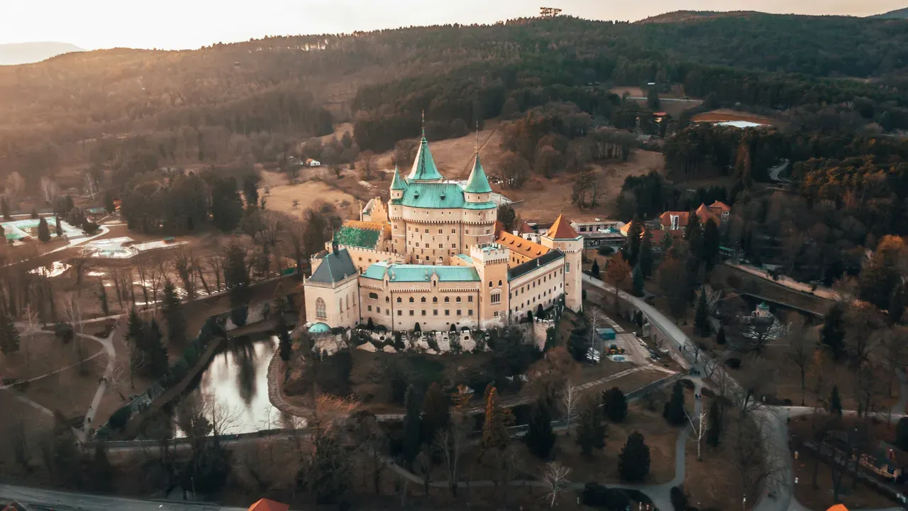 Aerial view of Bojnice Castle, a romantic castle with turquoise conical roofs, on a wooded hill above its moat in Slovakia at golden hour
