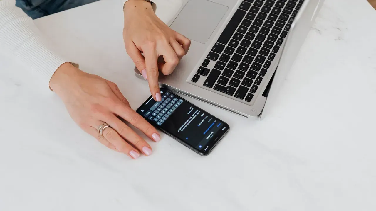 Close-up of hands using a smartphone next to a laptop on a white desk