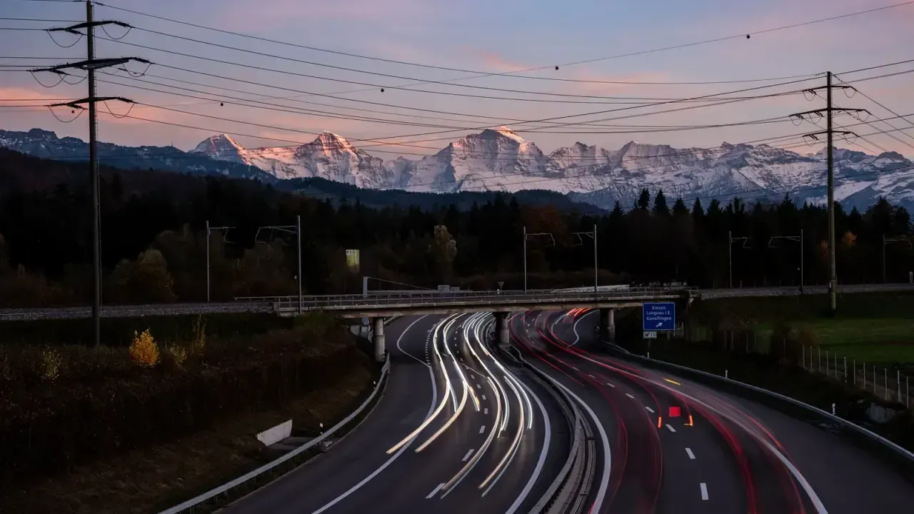 A Swiss motorway at dusk with light trails and snowy Alps in the background